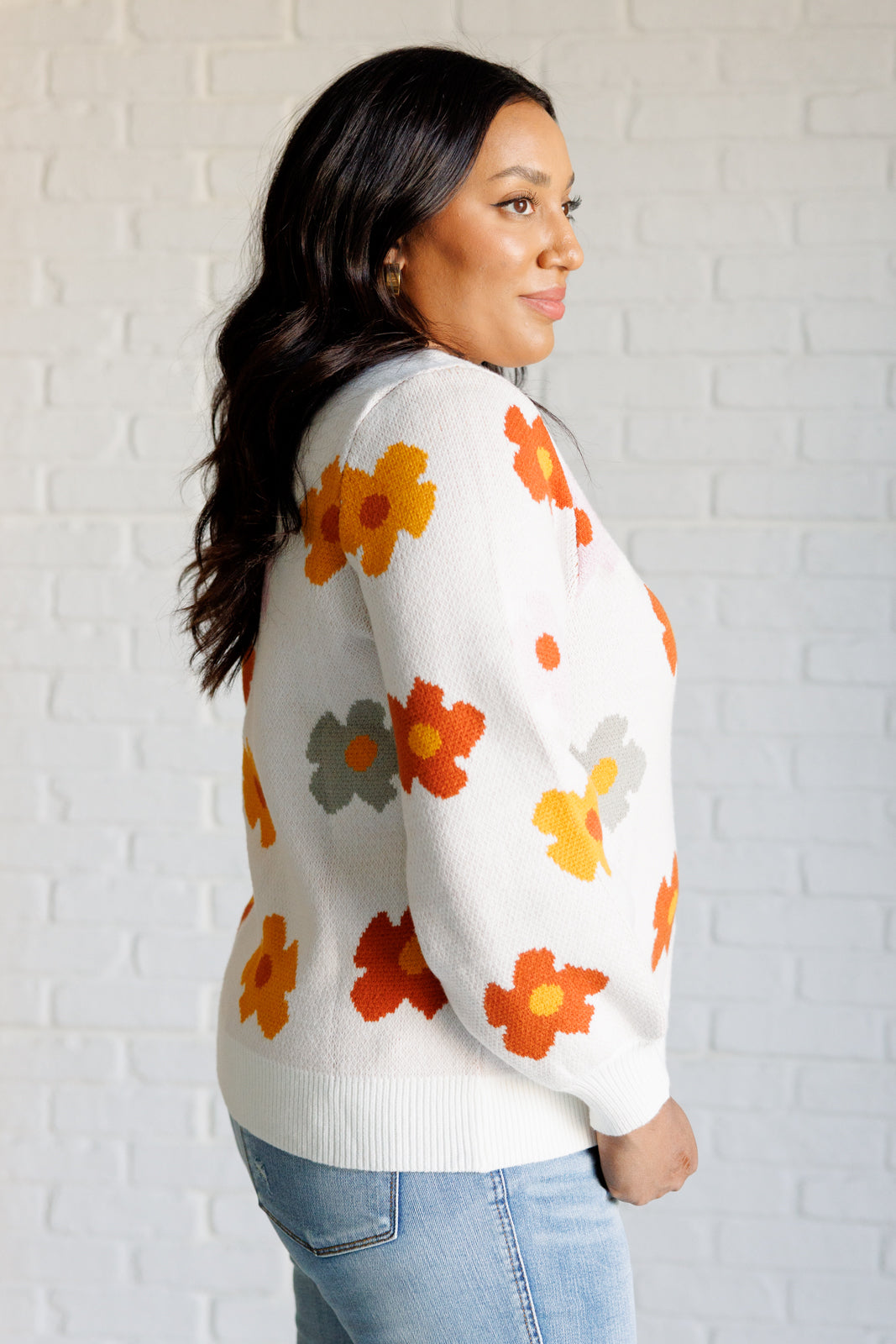 Woman wearing a white sweater with orange floral patterns against a light brick wall.