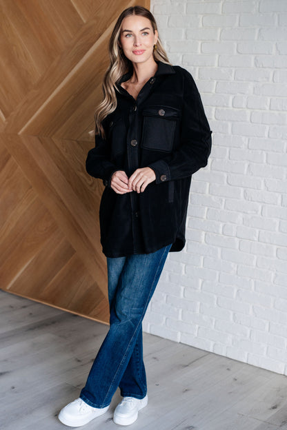 Woman wearing a black coat and blue jeans standing against a white brick wall.