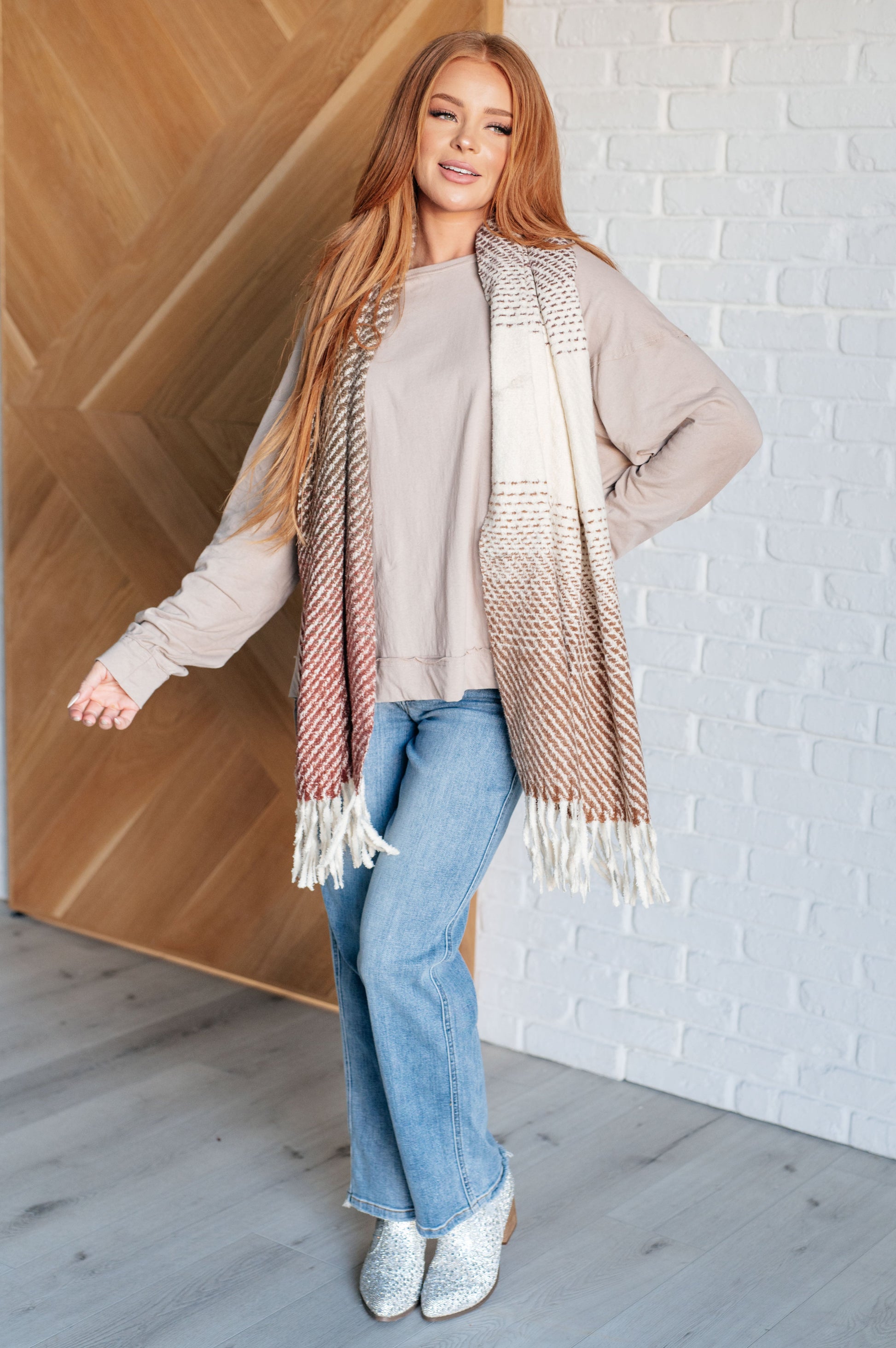 Woman wearing a beige sweater, blue jeans, and a patterned scarf against a white brick wall.