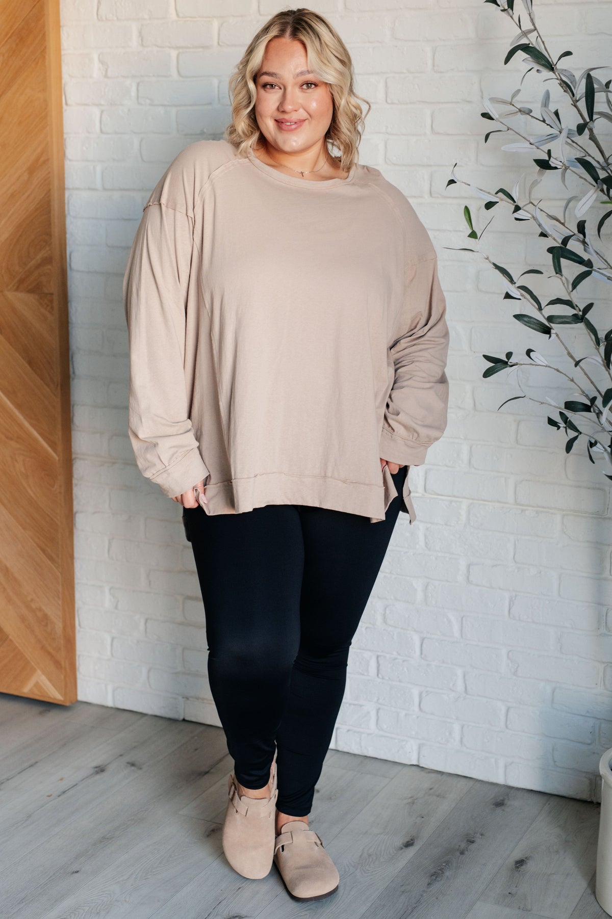 Woman wearing a beige sweater and black leggings standing against a white brick wall.