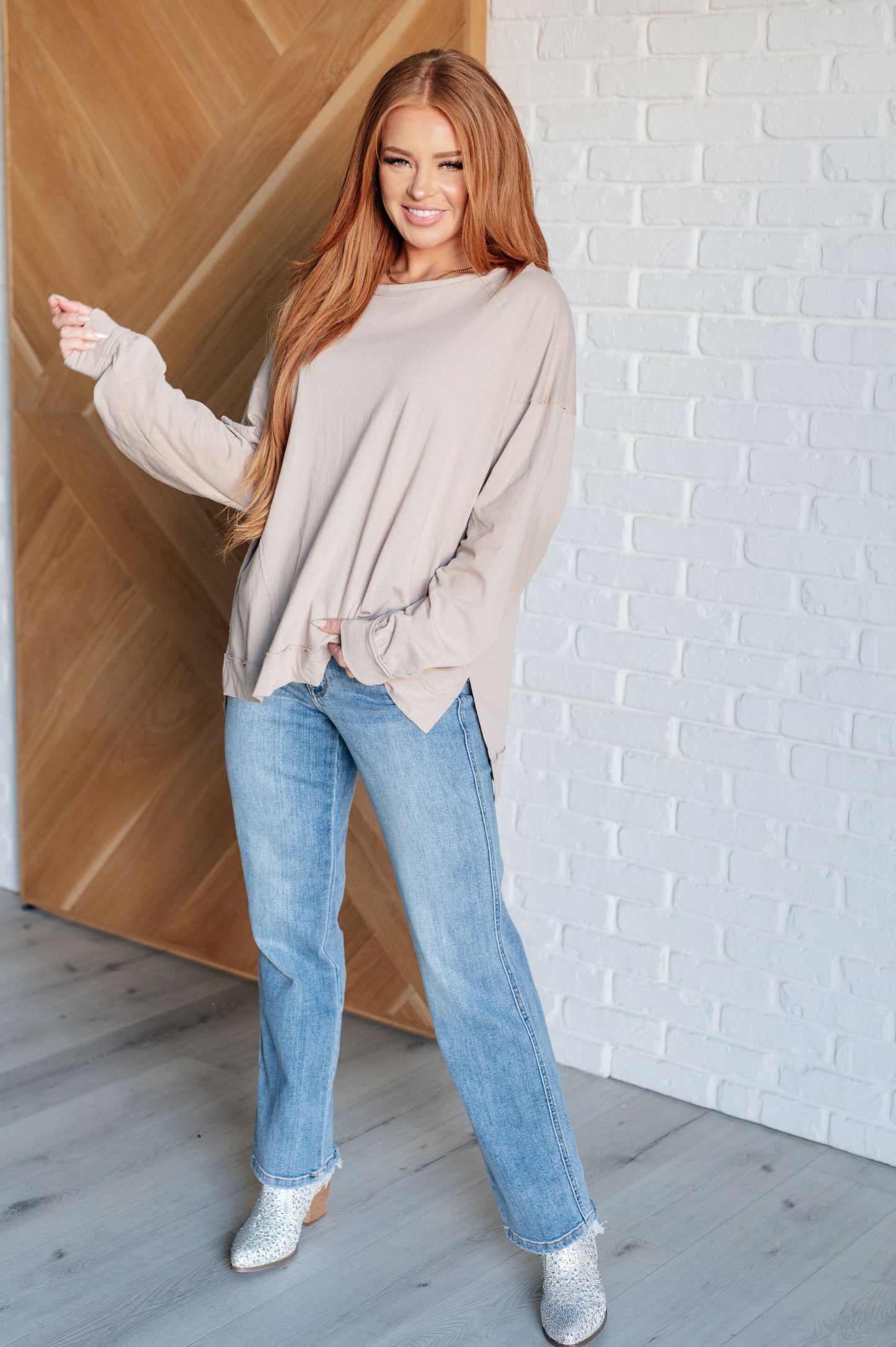 Woman wearing a beige sweater and blue jeans standing in a room with wooden and white brick walls.