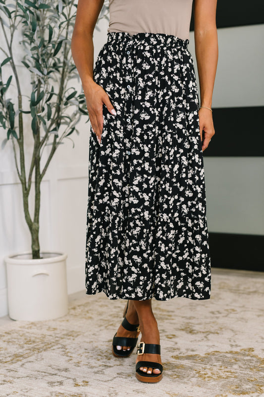 Person wearing a black floral skirt standing indoors with a plant in the background