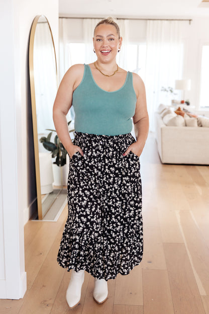 Woman wearing a green tank top and black floral skirt standing in a room with wooden flooring.