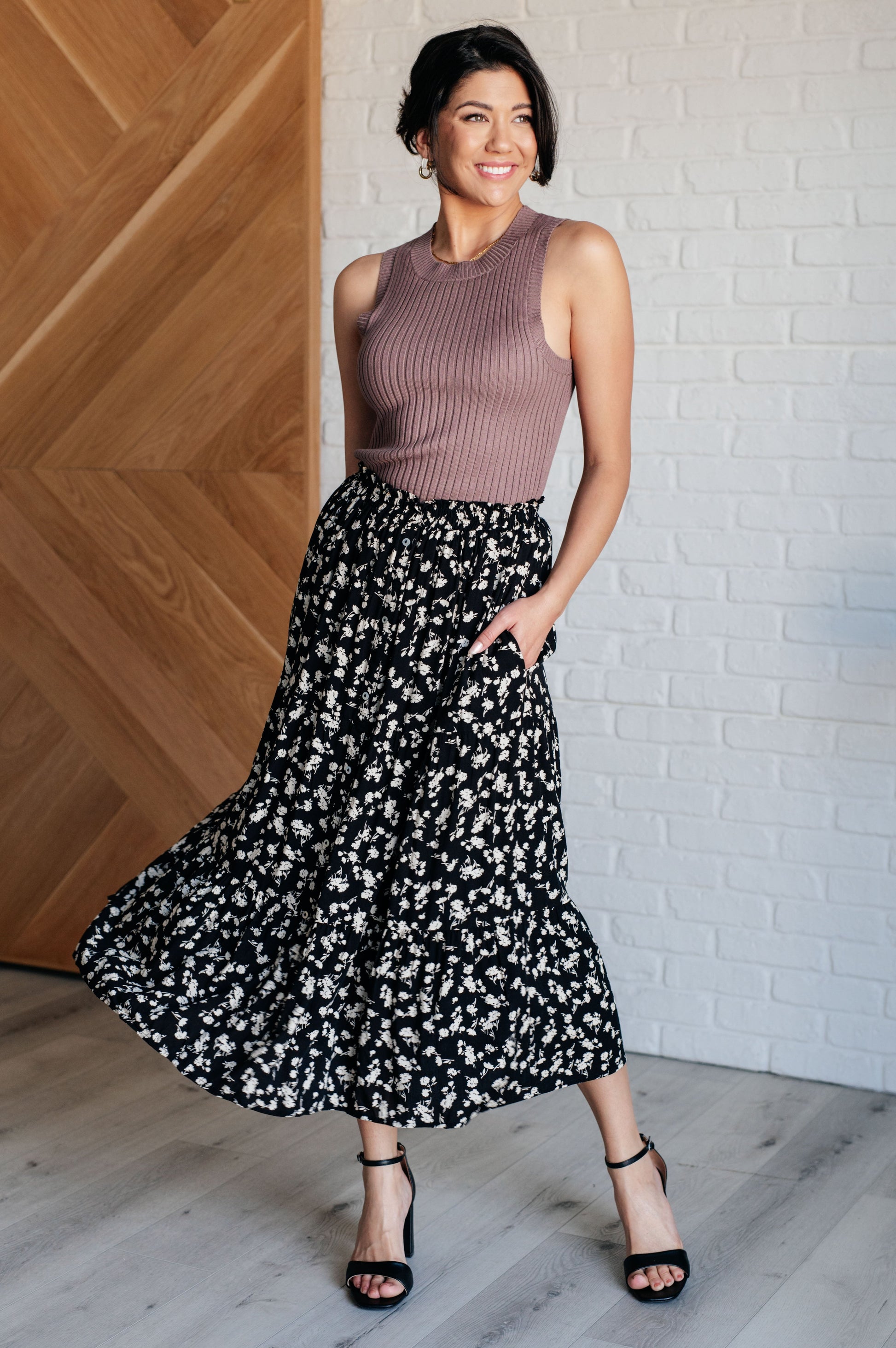 Woman wearing a sleeveless brown top and black floral skirt standing against a wooden wall.