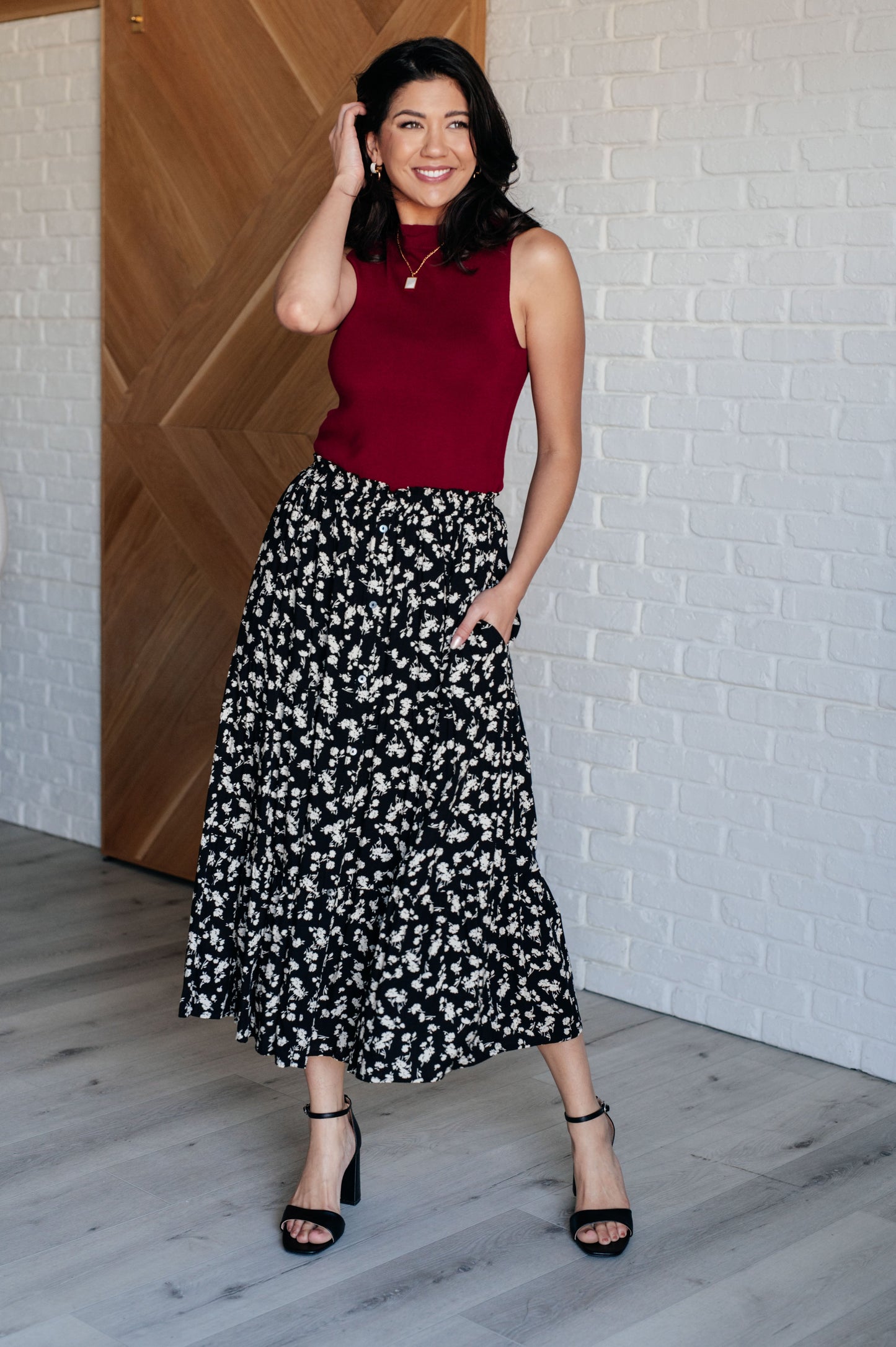 Woman wearing a red top and black floral skirt against a white brick wall.
