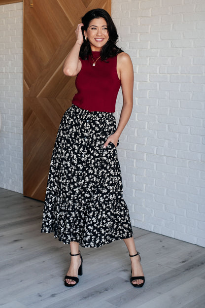 Woman wearing a red top and black floral skirt against a white brick wall.