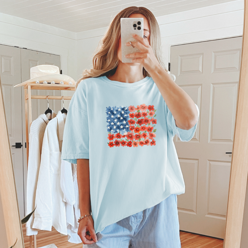 A person taking a mirror selfie wearing a light blue graphic tee with a floral flag design.