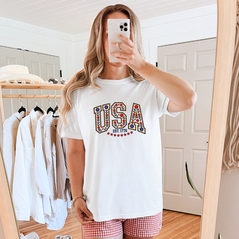 A person taking a mirror selfie wearing a white graphic tee with 'USA' multicolor text design and red and white checkered shorts.