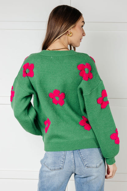 Green sweater with pink floral patterns worn by a person against a white wall.