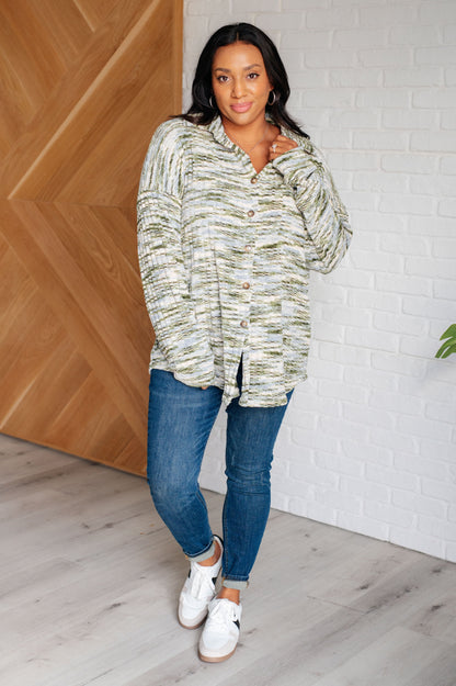 Woman wearing a patterned shirt and blue jeans standing indoors against a wooden wall.