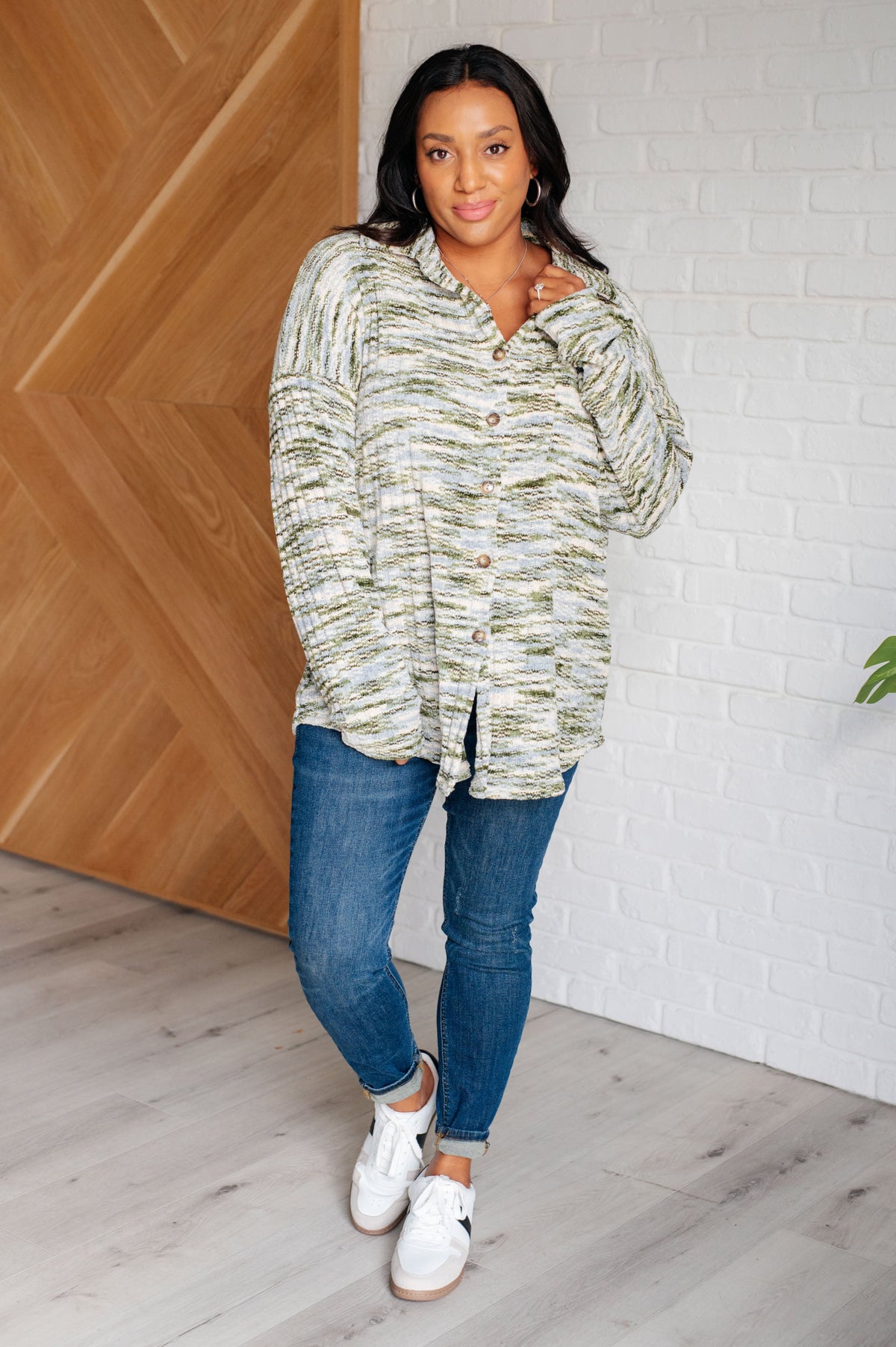 Woman wearing a patterned shirt and blue jeans standing indoors against a wooden wall.