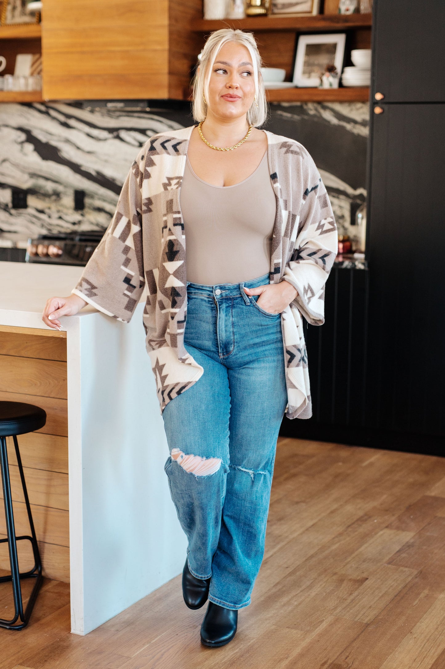 Woman standing in a kitchen wearing a patterned kimono, beige top, and blue jeans.