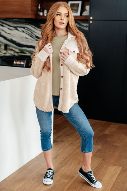 Woman wearing a beige jacket, green shirt, blue jeans, and black sneakers in a kitchen.
