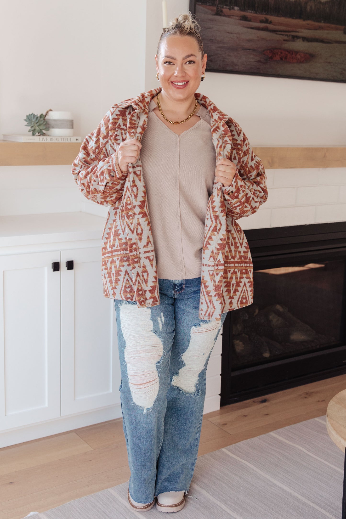 Woman wearing a patterned jacket, beige top, and ripped jeans in a living room.