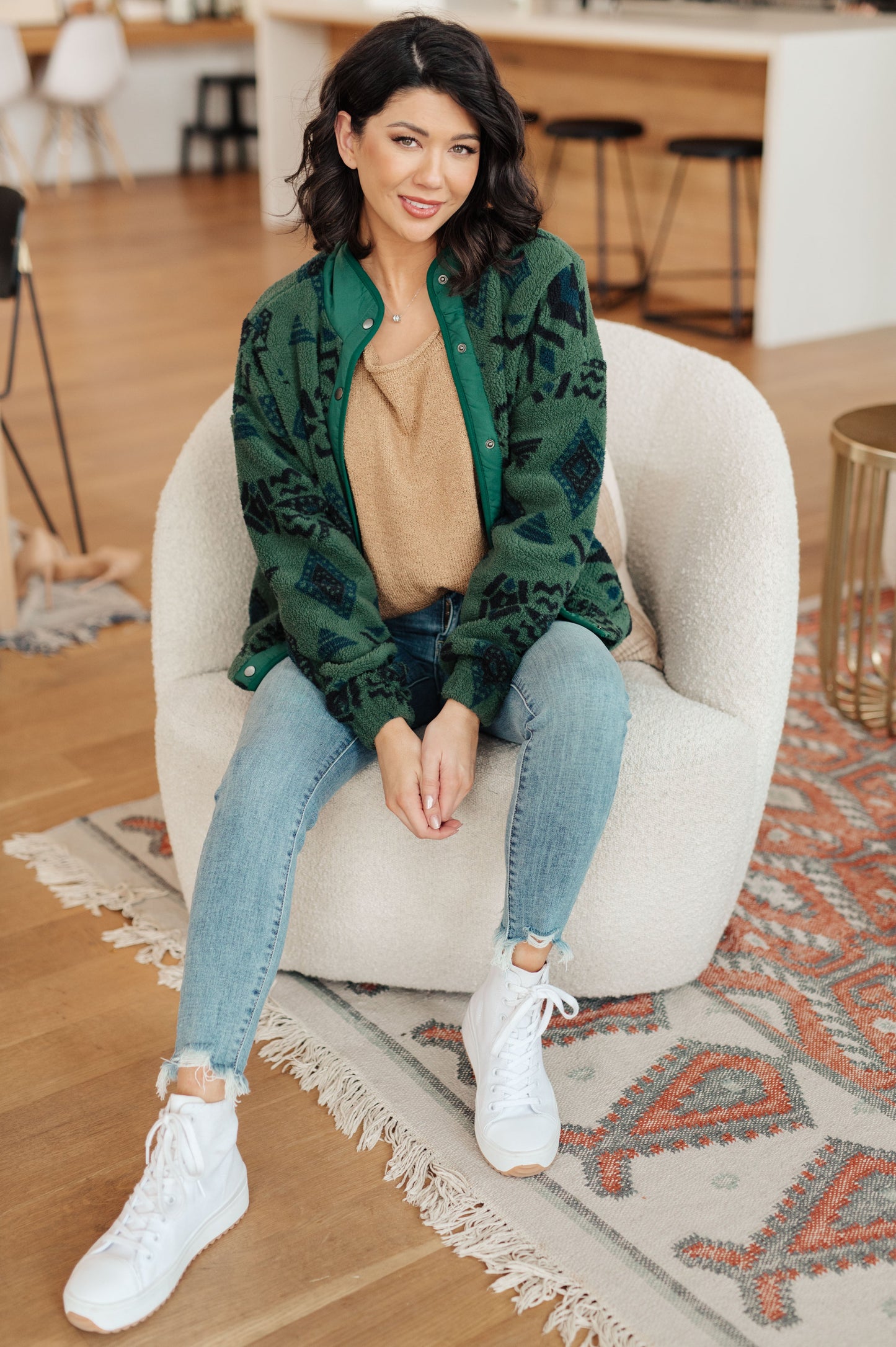 Woman sitting on a white chair wearing a green patterned jacket, beige sweater, blue jeans, and white sneakers in a room with wooden floor and white walls.