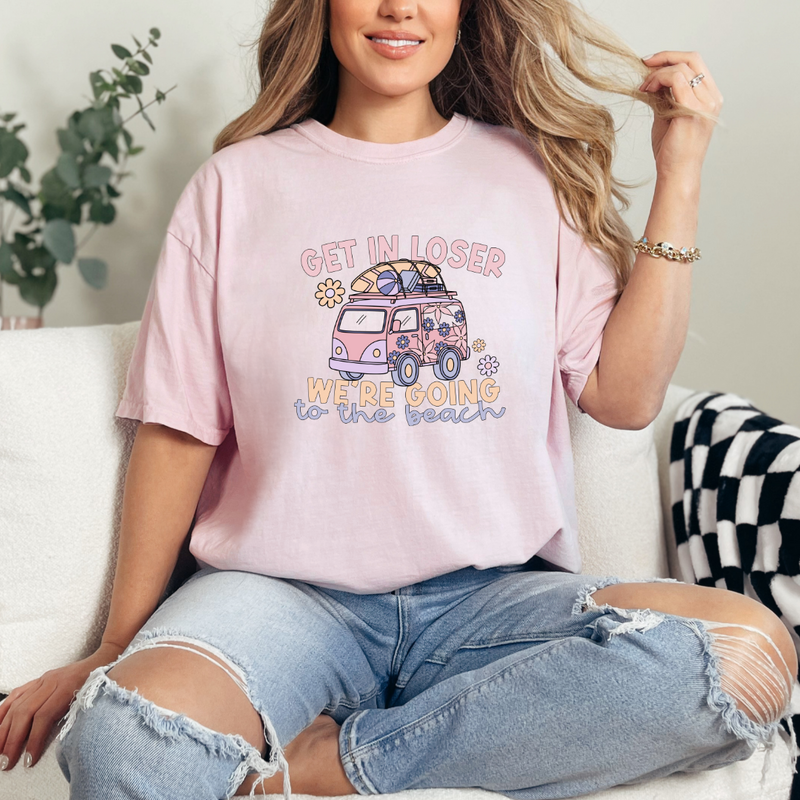 A person sitting on a couch wearing a pink graphic tee with the text 'Get in Loser We’re Going to the Beach' and a graphic of a vintage camper.