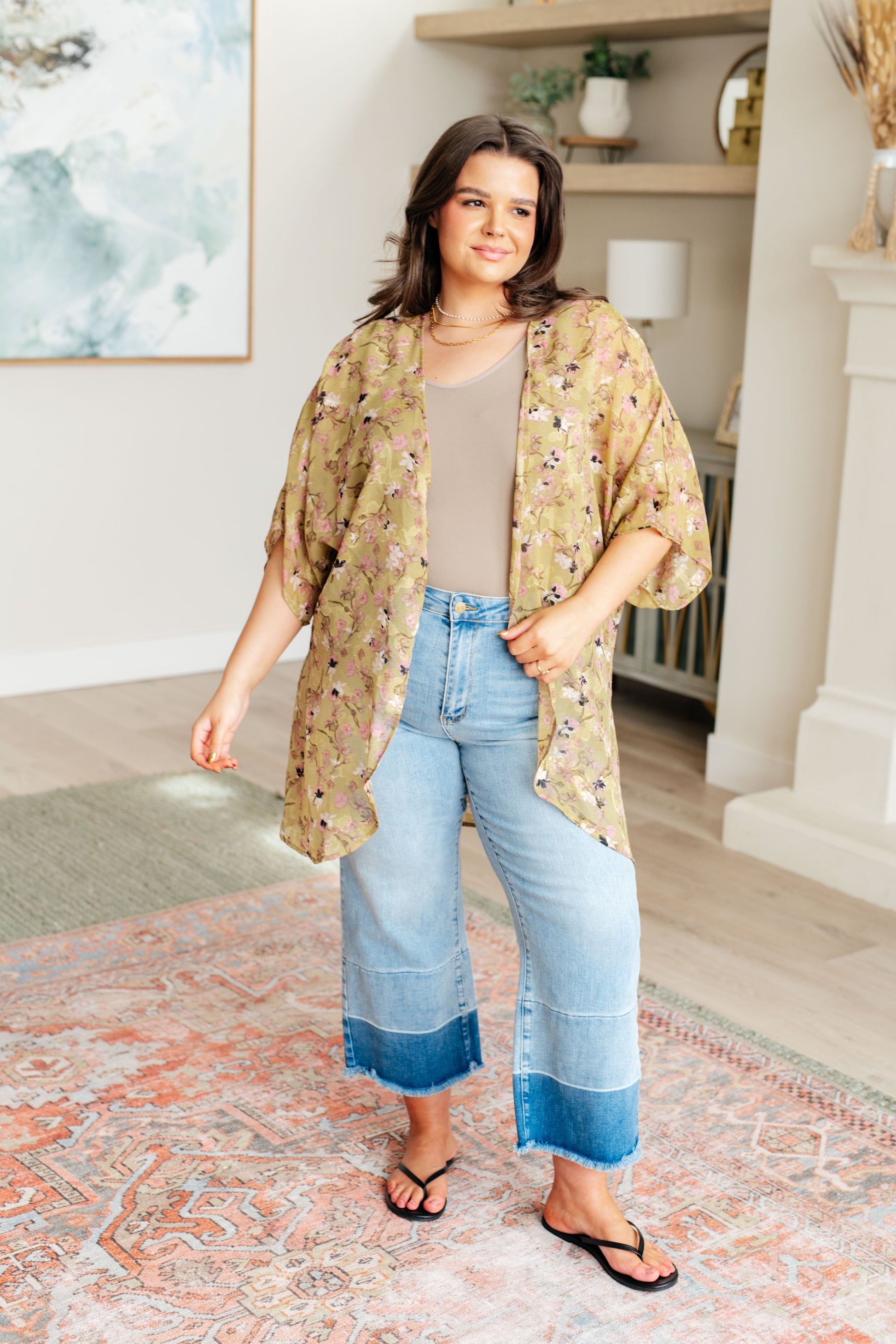 Woman wearing a floral kimono over a beige top and blue jeans in a home setting.