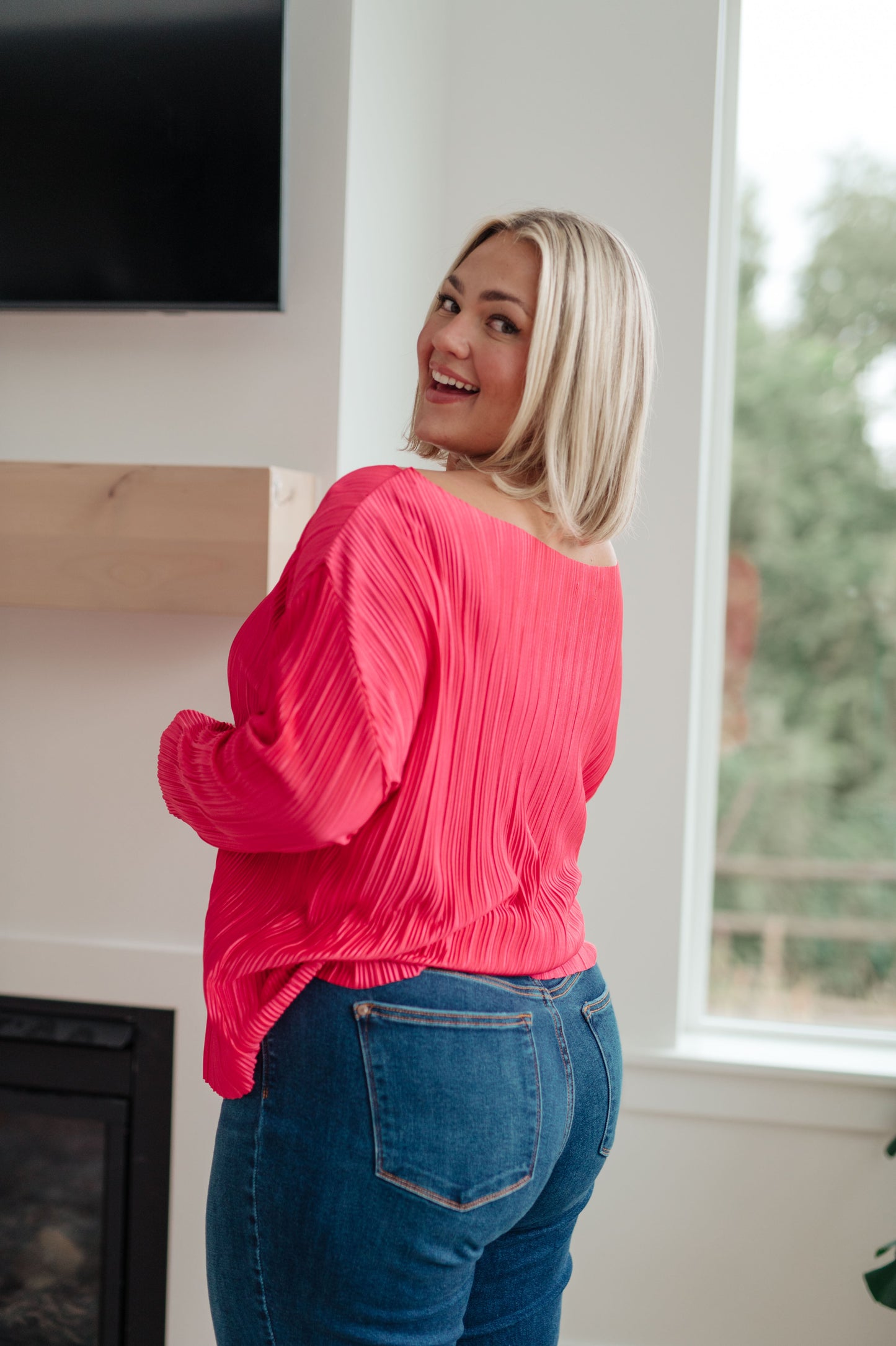 Woman wearing a pink blouse and blue jeans standing in a room with a fireplace and window.
