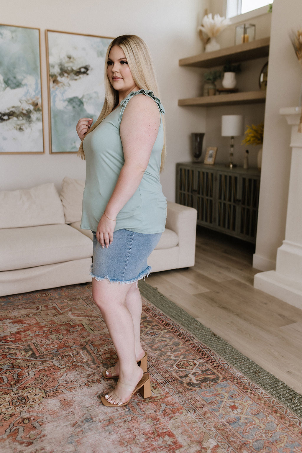 Woman standing in a living room wearing a light green top and denim shorts.