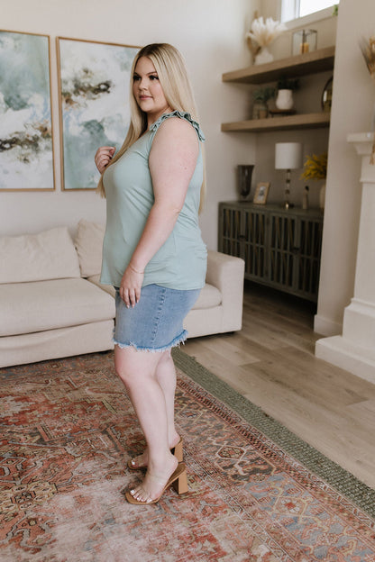 Woman standing in a living room wearing a light green top and denim shorts.