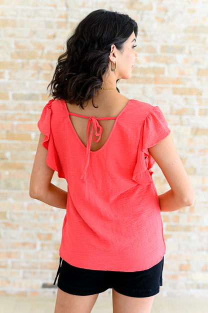 Woman wearing a coral top with ruffled sleeves and black shorts against a textured wall.