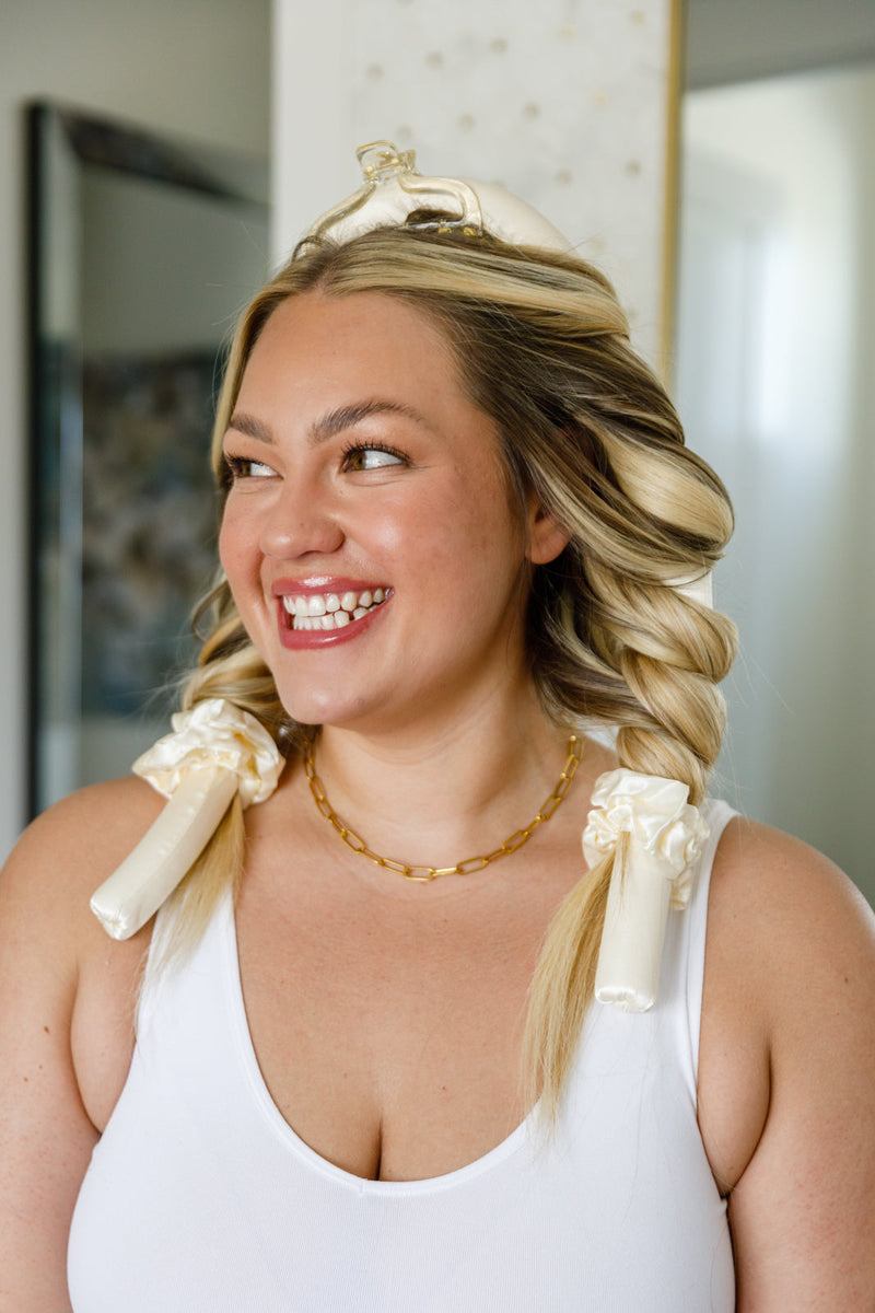 Woman with styled hair using hair ties, wearing a white tank top indoors.