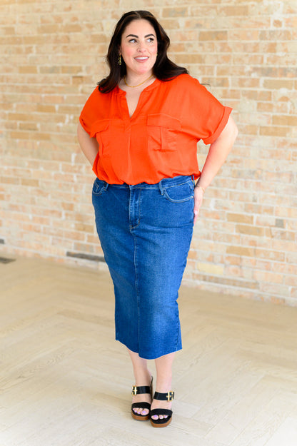 Woman wearing an orange blouse and blue jeans standing against a brick wall.
