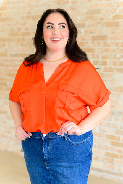 Woman wearing an orange blouse and blue jeans against a beige wall.