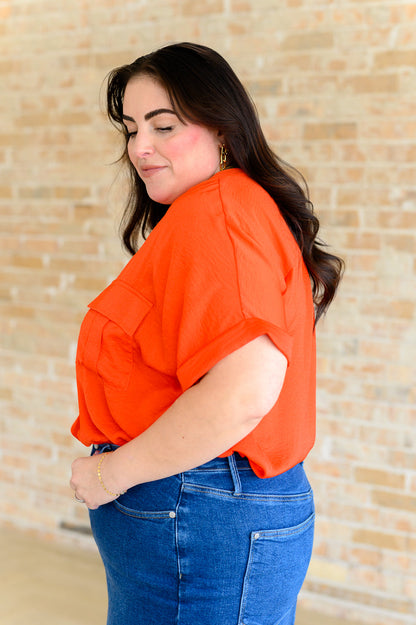 Woman wearing an orange blouse and blue jeans against a beige brick wall.