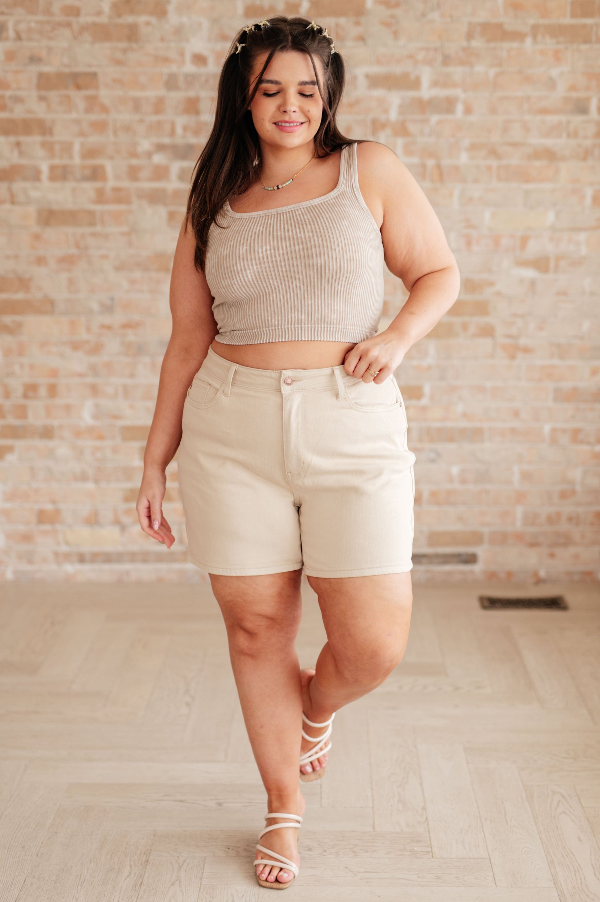 A woman is standing in a room wearing beige high-rise garment dyed shorts and a gray sleeveless 