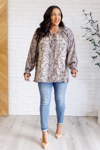Woman wearing a patterned blouse and blue jeans standing against a white brick wall.