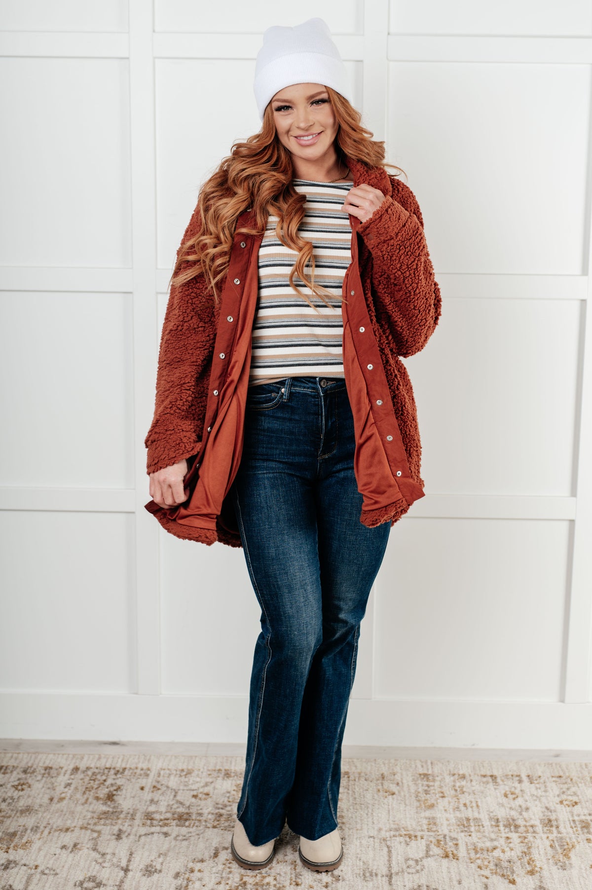 Woman wearing a rust-colored cardigan, striped shirt, and jeans against a white paneled wall.