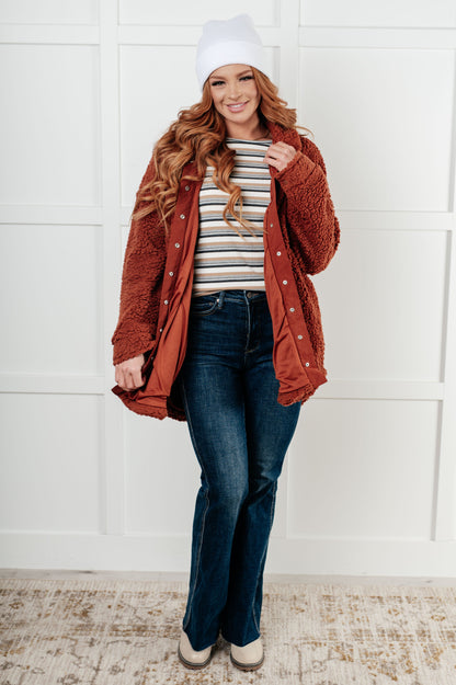 Woman wearing a rust-colored cardigan, striped shirt, and jeans against a white paneled wall.