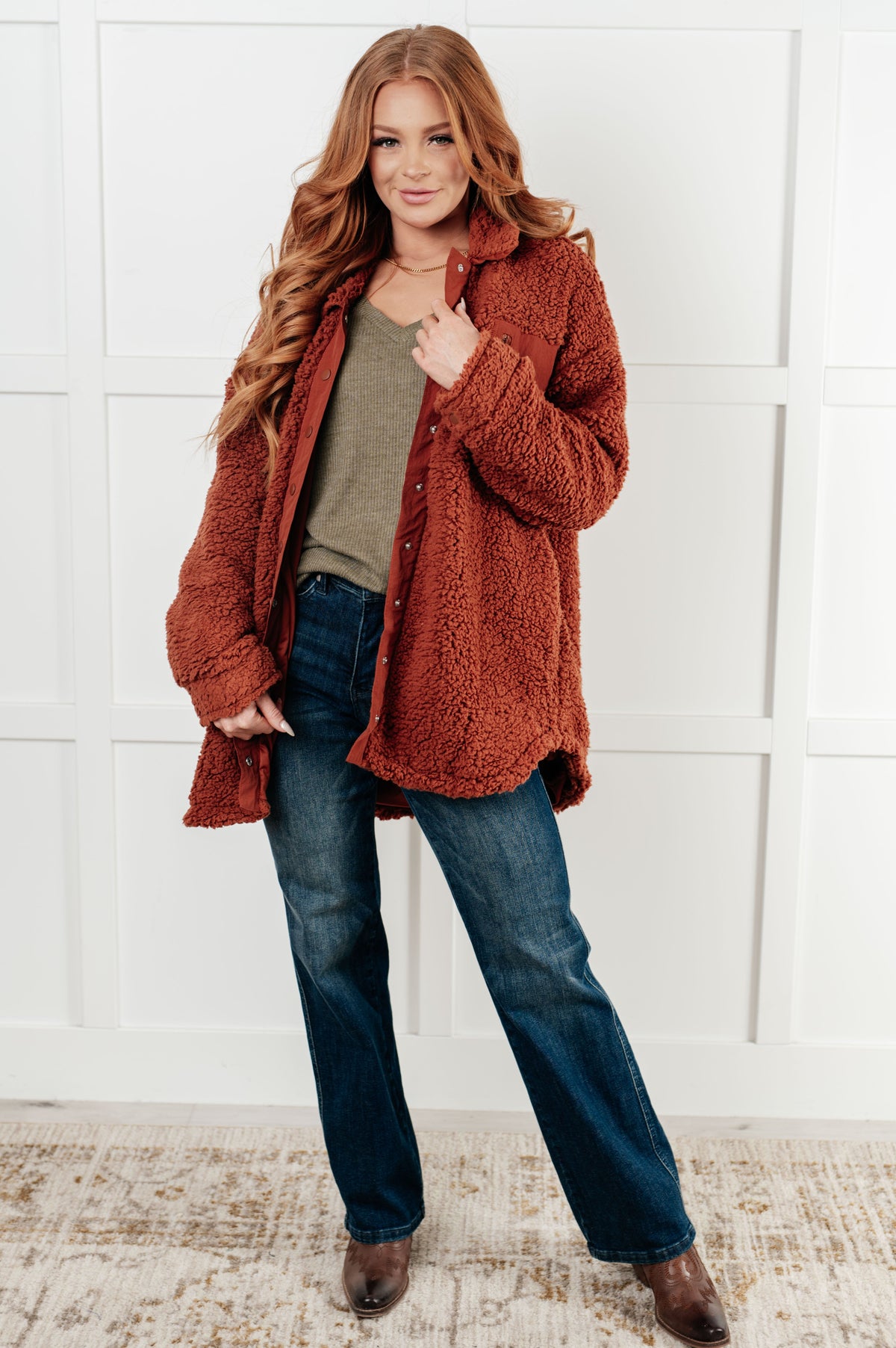 Woman wearing a rust-colored coat, green shirt, and blue jeans against a white wall.