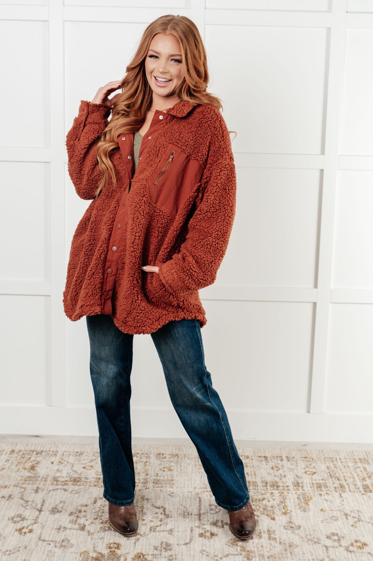 Woman wearing a brown sherpa jacket and jeans against a white wall.