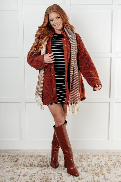 Woman wearing a rust-colored cardigan with fringe, striped dress, and brown knee-high boots against a white paneled wall.
