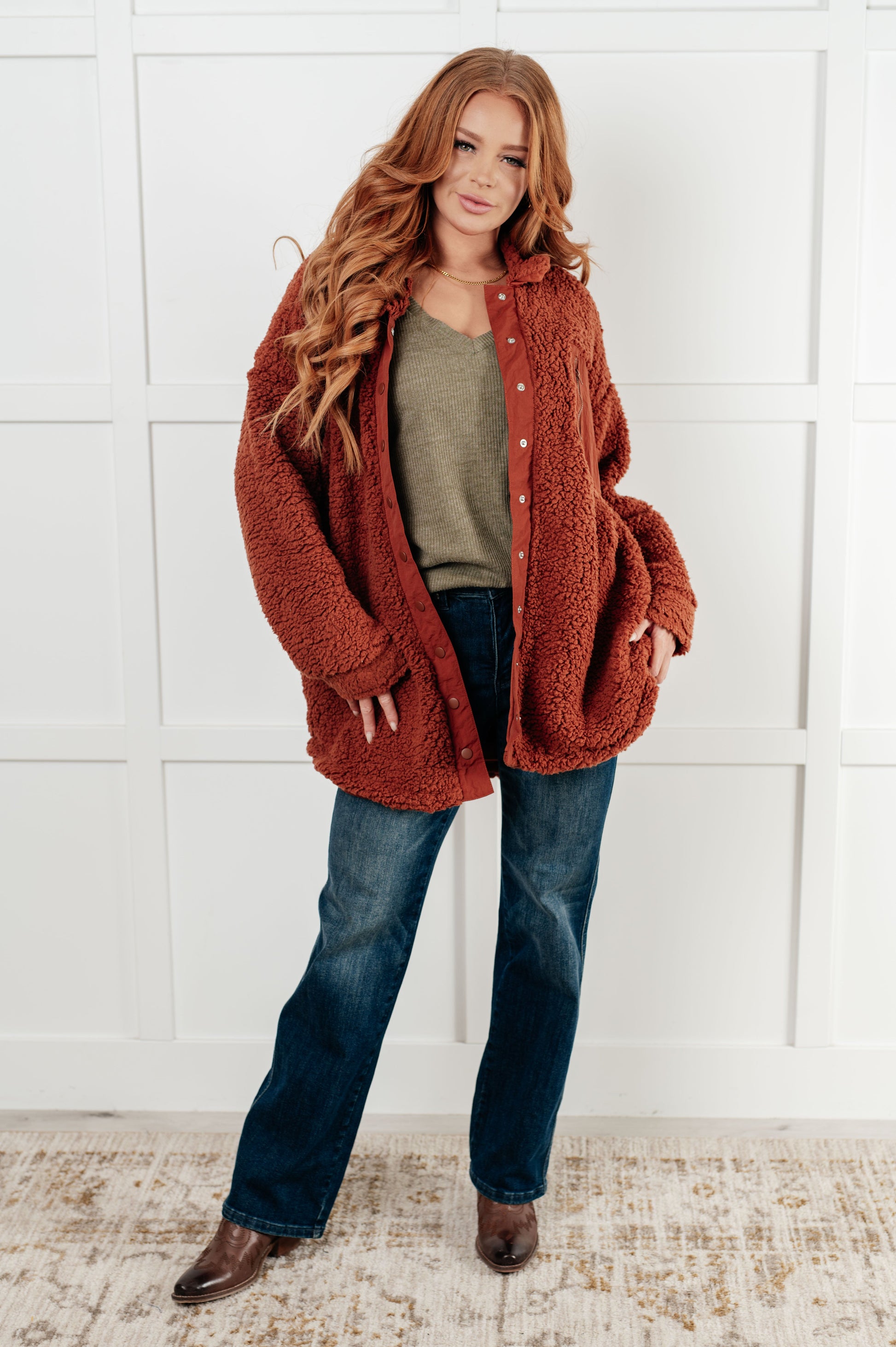 Woman wearing a rust-colored cardigan over a green shirt and blue jeans against a white wall.
