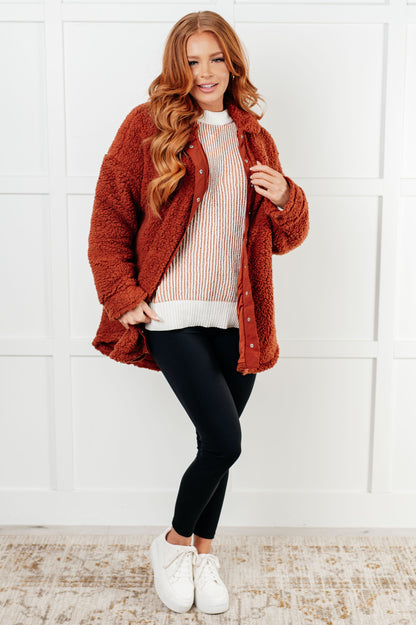Woman wearing a rust-colored fuzzy jacket over a white sweater with black pants and white shoes on a white background