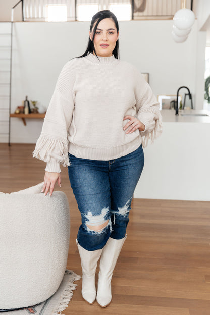 Woman wearing a beige sweater, blue jeans, and white boots in a kitchen.