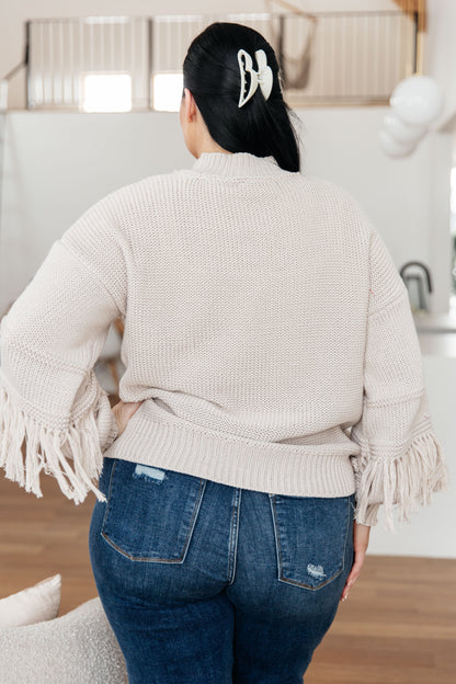 Person wearing a beige sweater with fringe details and blue jeans in an indoor setting.