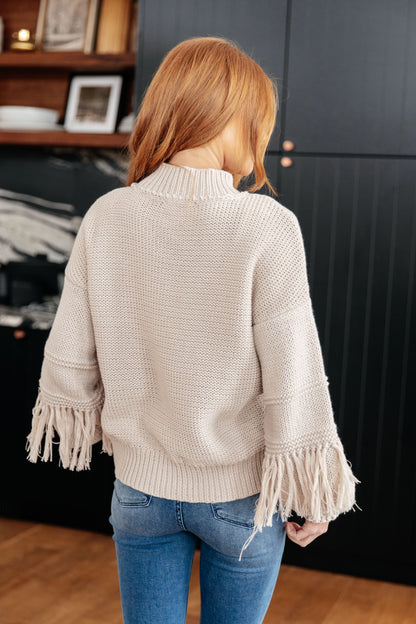 Person wearing a beige sweater with fringe details in an indoor setting
