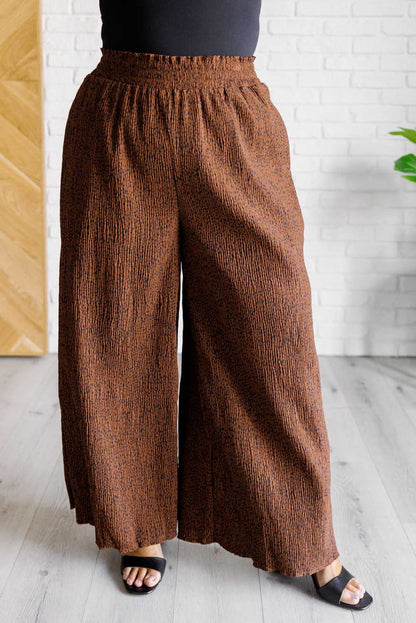 Brown textured wide-leg pants worn by a person against a white brick wall.