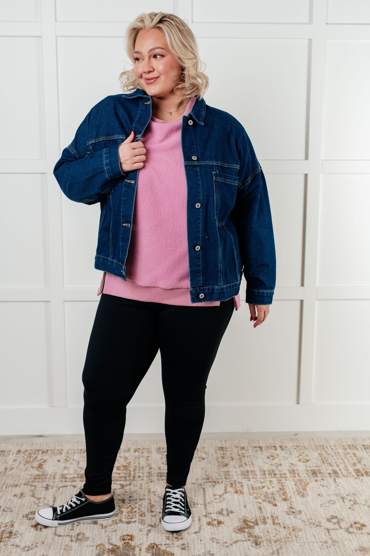 Woman wearing a denim jacket, pink shirt, black pants, and black sneakers against a white wall.