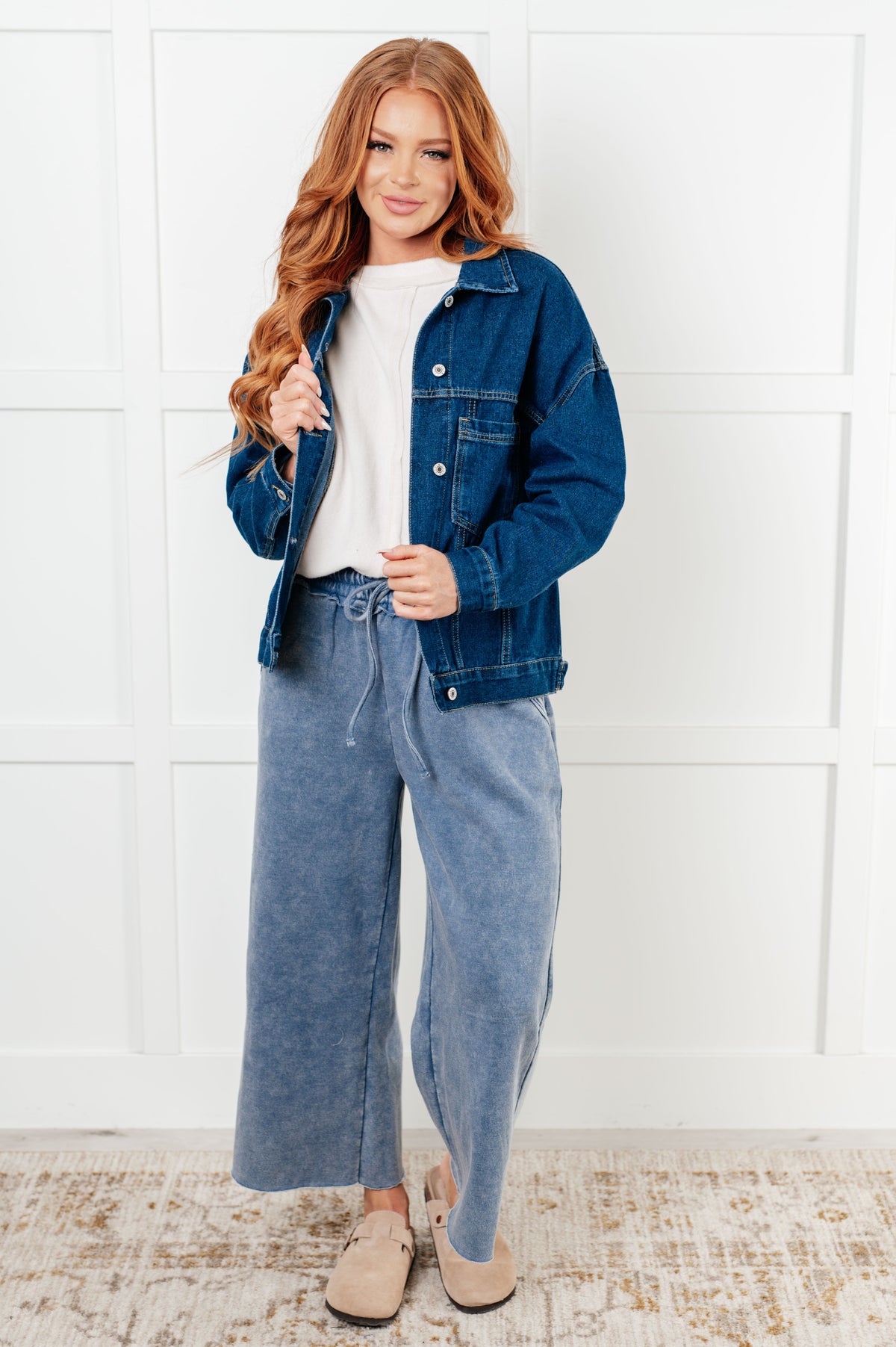 Woman wearing a blue denim jacket and wide-leg jeans against a white wall.
