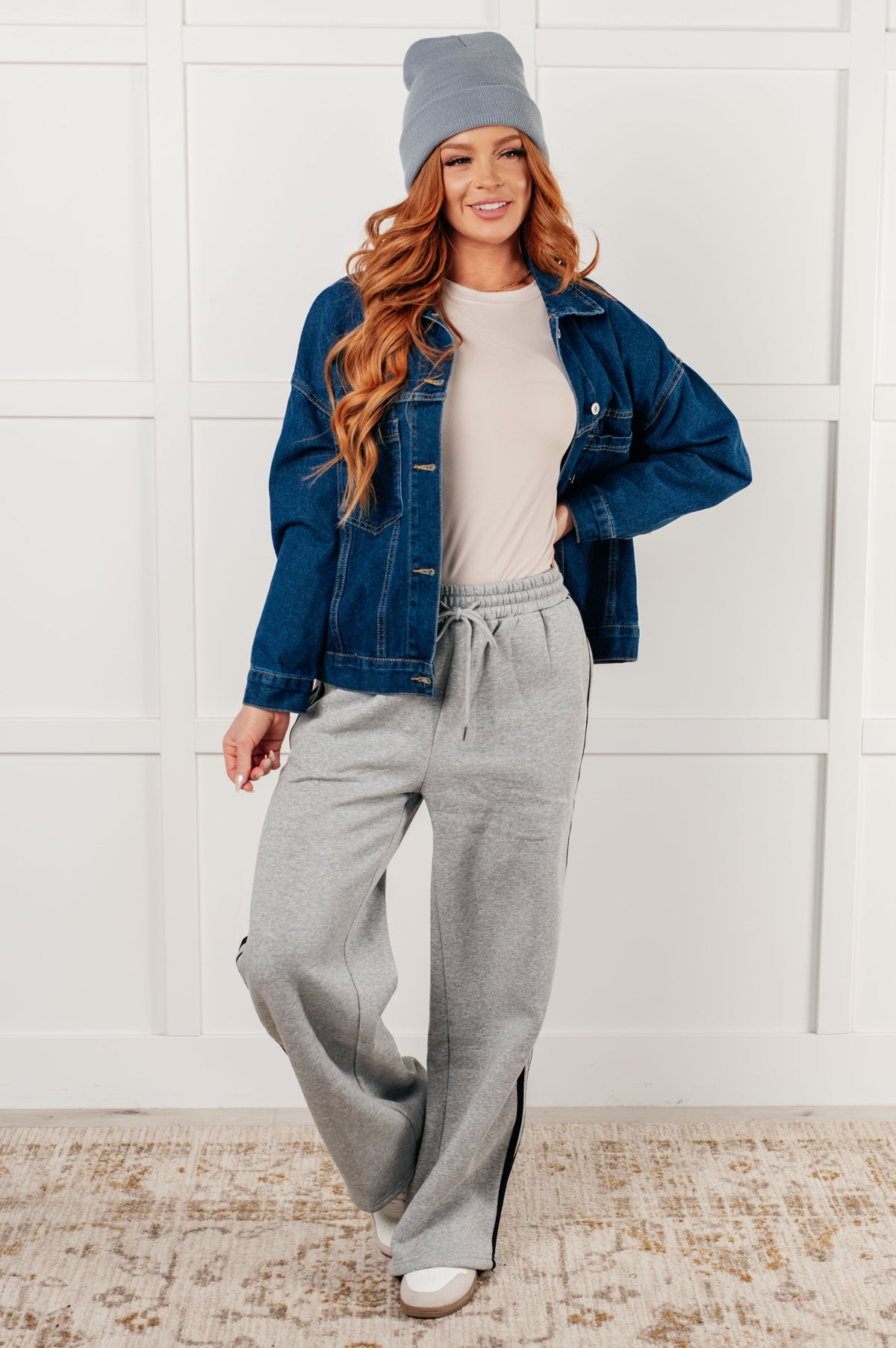 Woman wearing a denim jacket, gray beanie, and gray sweatpants against a white wall.