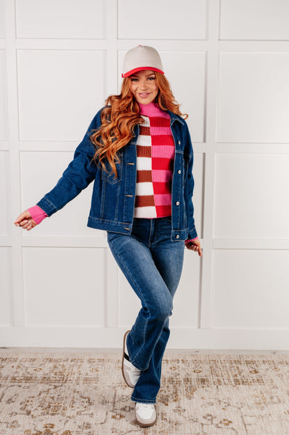 Woman wearing a denim jacket, checkered sweater, and jeans against a white wall.
