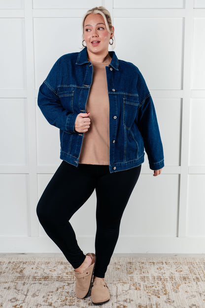 Woman wearing a denim jacket, beige top, black leggings, and brown shoes against a white paneled wall.