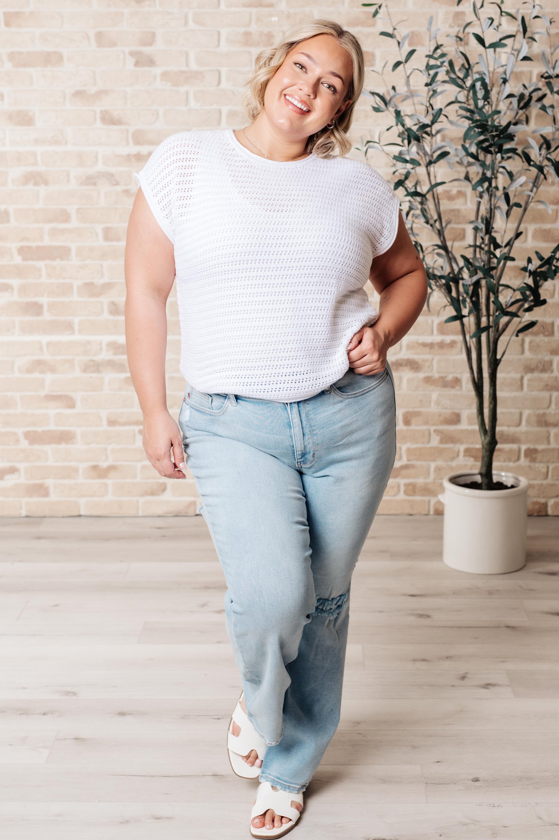 Woman wearing a white knit top and light blue jeans standing in a room with a plant and brick wall.