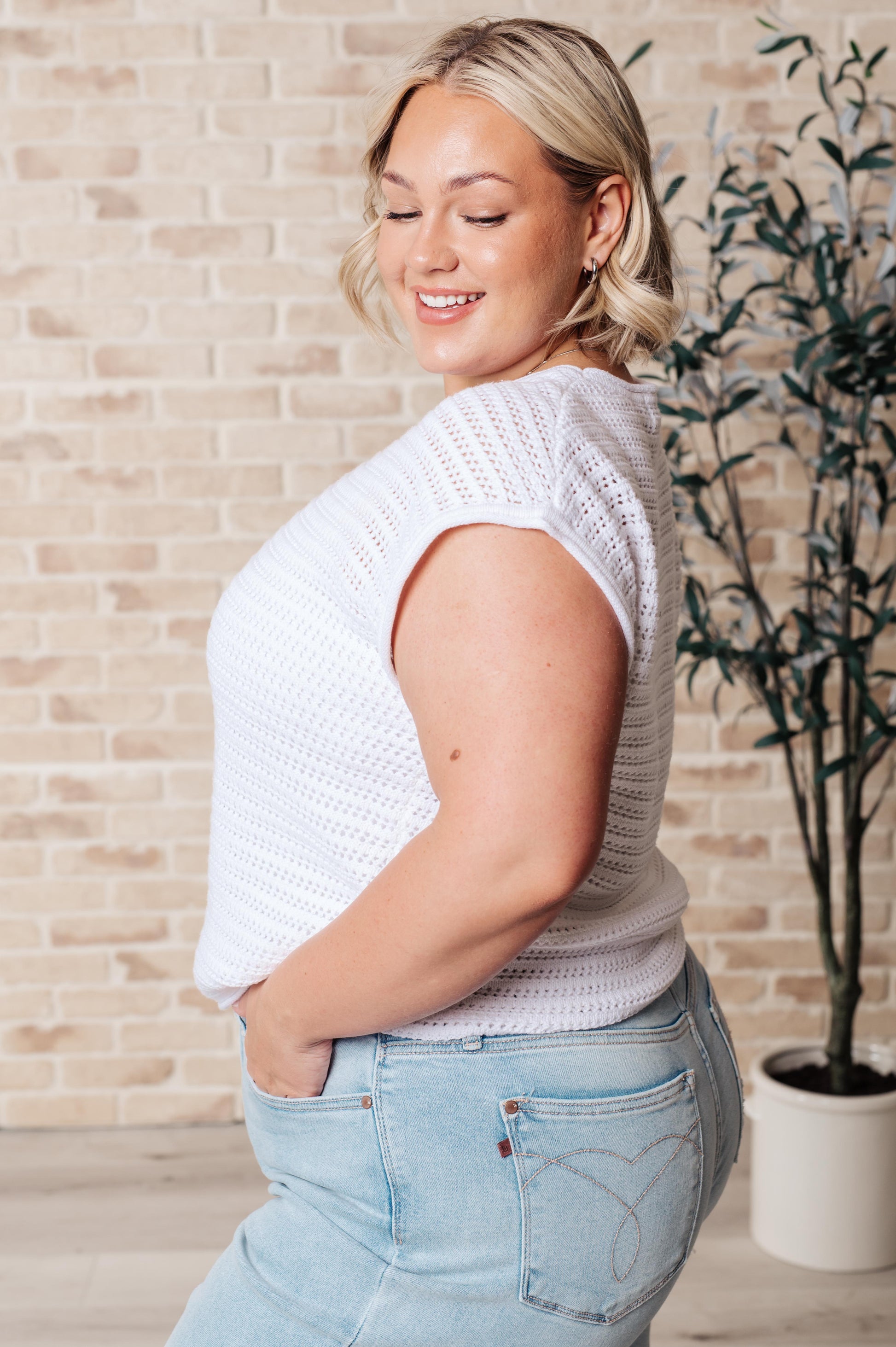Woman wearing a white knit top and light blue jeans standing against a brick wall with a plant.