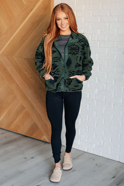 Woman wearing a green patterned jacket and black leggings standing against a white brick wall.